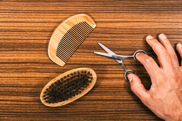 Hand holding small scissors to cut your hair and trim your beard yourself with a modern wooden comb and beard brush. Hair Tools for Men\'s Haircut on a vintage wooden base.
