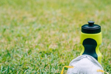 Yeşil çimen parkında maskeyle oynanacak bir su şişesi. Koronavirüsü önlemek için koruyucu önlemler alarak dışarıda spor yapmak. Kopyalama alanı için yeşil çimen arkaplanı.