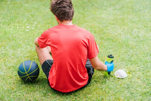 Sporting boy practicing outdoor sports with protective measures to prevent the spread of the coronavirus. Person with a basketball, a mask and a water bottle sitting on the park\'s grass.