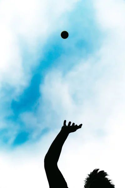 Happy person doing sport by throwing a ball in the air. Silhouette of a carefree boy playing happily with a tennis ball on a beautiful sunny summer day.