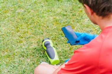 Parktaki yeşil çimlerde oturan çocuk, açık hava sporları için gerekli koronavirüs önleme önlemleriyle birlikte mavi lateks eldivenli cep telefonuna danışıyor..