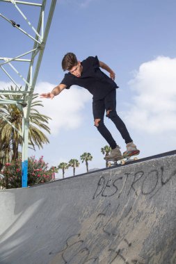Genç kaykaycı, sokak arkaplanı ve mavi gökyüzü ile parkta numaralar yapıyor..