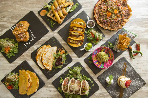 Yukarıdan görülen tipik Meksika yemekleriyle dolu bir masa. Nacho, taco, chimichanga, quesadilla, guacamole.