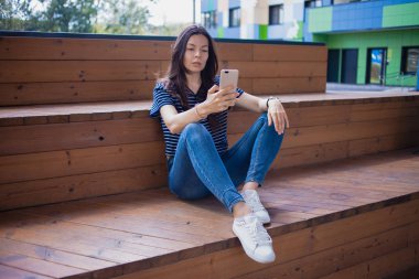 Esmer bir kız, büyük kol saati olan bir öğrenci, tahta basamaklarda bacaklarıyla oturur ve akıllı telefonu dikkatle kontrol eder..