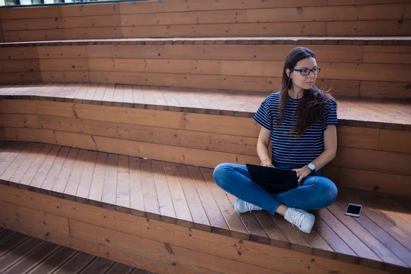 Una seria studentessa bruna, si siede su gradini di legno, con un computer portatile aperto, digitando su una tastiera, impara online . — Foto Stock