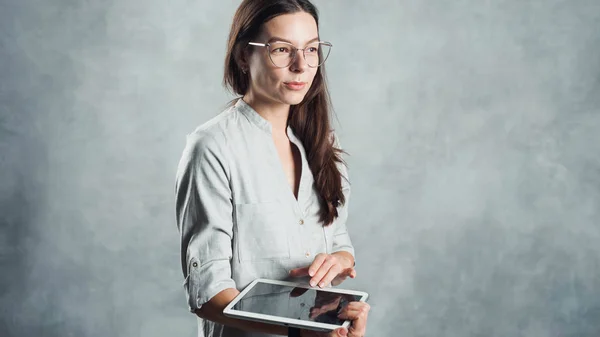Jonge succesvolle zelfverzekerde vrouw in een grijs shirt maakt gebruik van een digitale tablet voor werk en toegang tot internet — Stockfoto