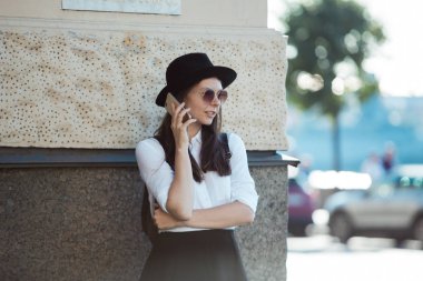 şapkalı ve beyaz gömlekli genç kadın şehirde yürür ve akıllı telefon kullanır.