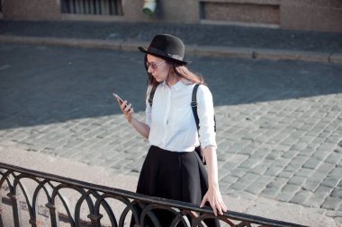 bir şapkalı genç kadın ve bir sırt çantası ile şehirde yürür ve bir akıllı telefon kullanır.