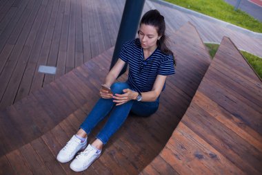 Esmer bir kız, uzun saçlı, geniş kol saati olan, kot pantolonlu, ahşap basamaklarda oturuyor ve akıllı telefonu kontrol ediyor..
