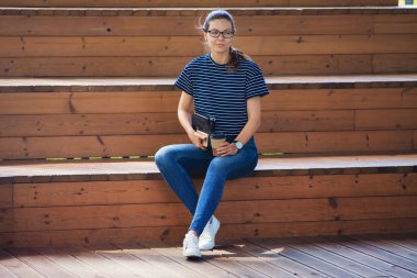 Hoş bir genç kız elinde bir fincan kahve ve dizüstü bilgisayarla ahşap basamaklarda oturuyor, işini bitirip mola veriyor..