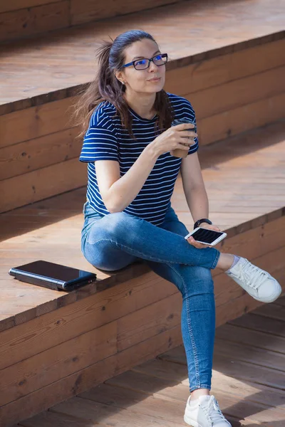 Young girl is sitting on steps, finished work, smiles, checks the phone, drinks coffee.