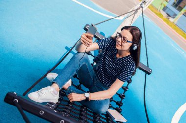 Kız, bir esmer, bir hamakta oturuyor, sways, müzik dinlerken, dans, rahatlatıcı, şarkı, bir WiFi yakalamak için çalışıyor.