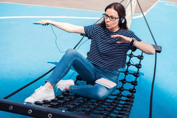 Meisje, een brunette, zit in een hangmat, sways, luisteren naar muziek, dansen, ontspannen, zingen, proberen om een WiFi te vangen. — Stockfoto