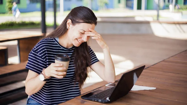 Een brunette meisje zit achter houten bureau, kijkt naar een laptop monitor, glimlacht, lacht, praat online, koffie drinken, werkt. — Stockfoto