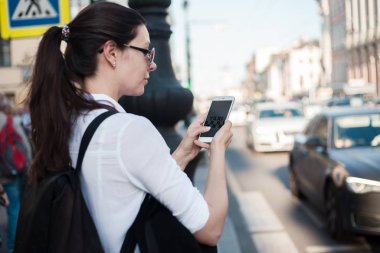 Mobil uygulamayı kullanarak taksi çağırın. Şehirde genç bir kadın elinde bir akıllı telefon ile yolun yanında duruyor