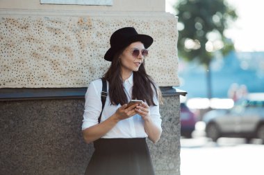 şapkalı ve beyaz gömlekli genç kadın şehirde yürür ve akıllı telefon kullanır.