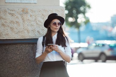 şapkalı ve beyaz gömlekli genç kadın şehirde yürür ve akıllı telefon kullanır.