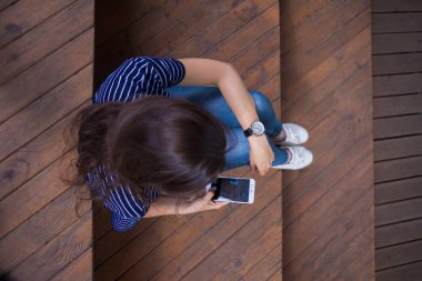 Büyük bir bilek saati ile esmer kız, ahşap adımlarla oturur ve bir akıllı telefon kontrol eder. Görünüm yukarıda açıkça.