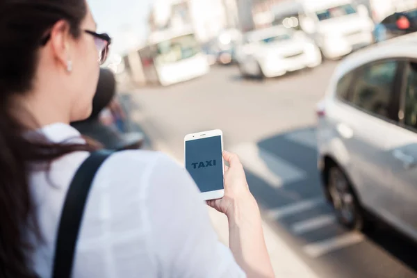 Call a taxi using the mobile application. A young woman in the city stands near the road with a smartphone in her hand