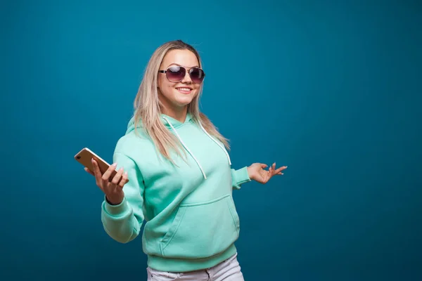 Rubia alegre con un teléfono móvil. Retrato de una joven mujer positiva con capucha azul — Foto de Stock