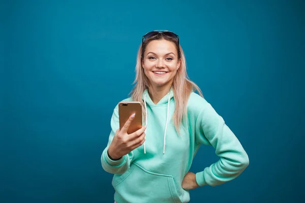 Loura alegre com um telefone celular. Retrato de uma jovem mulher positiva com capuz azul — Fotografia de Stock