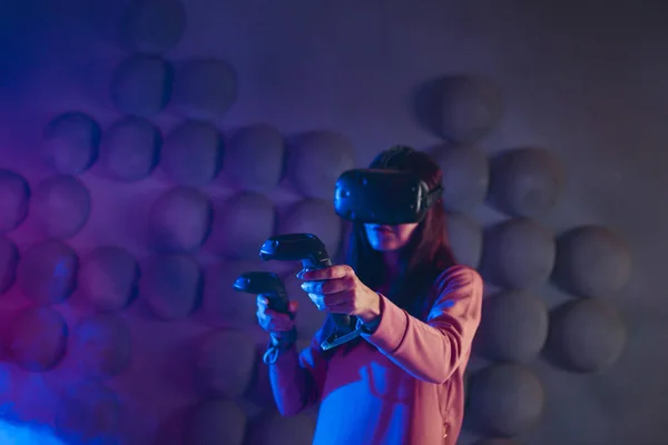 First-person shooter. A woman with glasses and a virtual reality helmet uses game controllers in both hands, like guns.