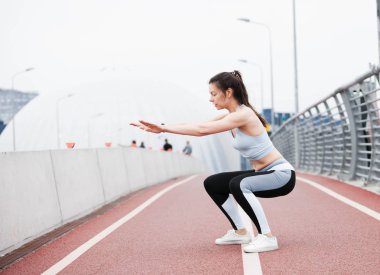 Spor elbiseli genç bir kadın şehir manzarası ve beyaz gökyüzüne karşı çömelir, hamleler yapar, spor yapar..