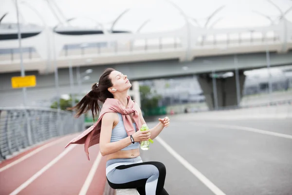 Sportowa kobieta w stroju sportowym po joggingu, pije wodę z jasnozielonej butelki, siedzi na parapecie mostu. — Zdjęcie stockowe