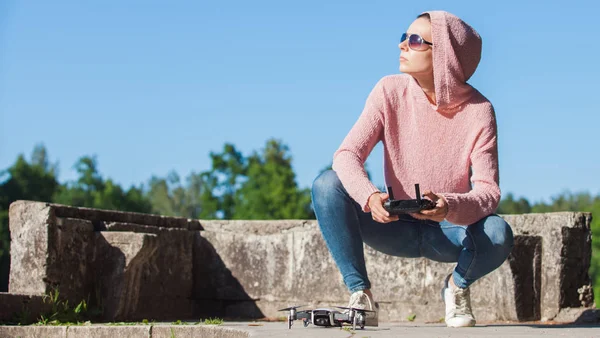 Een jonge vrouw in een roze capuchon en een zonnebril bestuurd met de afstandsbediening drone landing in de natuur. — Stockfoto