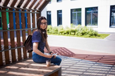 Genç bir kadın, bir öğrenci, gözlüklü bir esmer, bir bankta oturuyor, fotoğraf çekiyor, etrafa bakıyor..