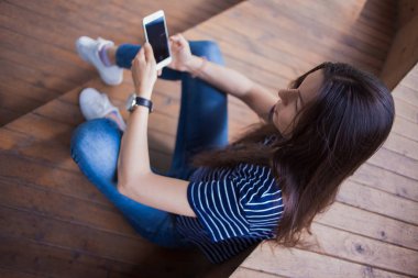 Büyük kol saati olan esmer bir kız, ahşap merdivenlerde oturur ve akıllı telefonu kontrol eder. Manzara yukarıdan..