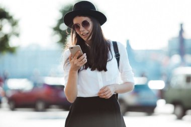 şapkalı ve beyaz gömlekli genç kadın şehirde yürür ve akıllı telefon kullanır.