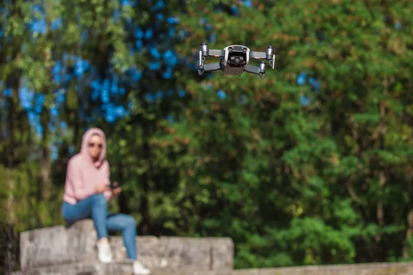 Een jonge vrouw in een roze hoodie en zonnebril zittend op een stenen Bank en bediend door afstandsbediening drone vlucht. — Stockfoto