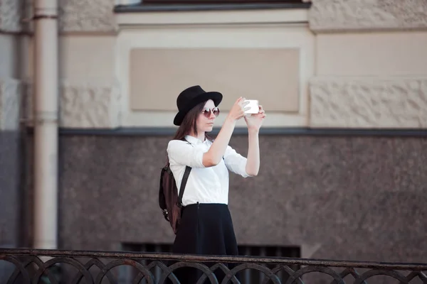 Toerist in de stad neemt een foto op de smartphone. Een jonge vrouw in een zwarte hoed en een wit overhemd, — Stockfoto