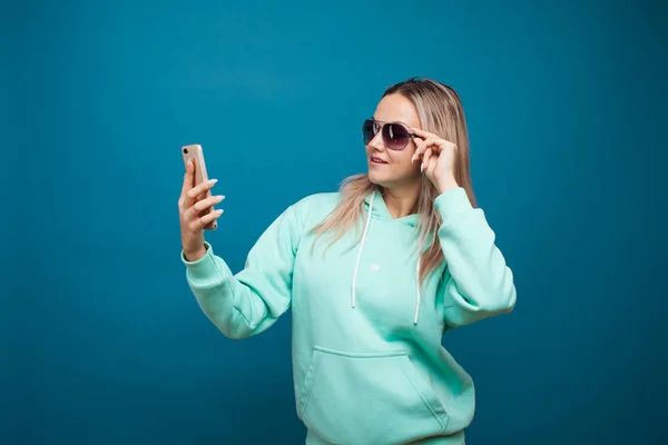 Elegante moderno hipster menina com capuz azul com o capuz usa um smartphone. — Fotografia de Stock