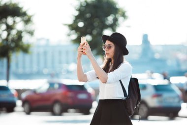 Şehirde turist Smartphone bir fotoğraf çeker. Siyah şapkalı genç bir kadın ve beyaz bir gömlek,
