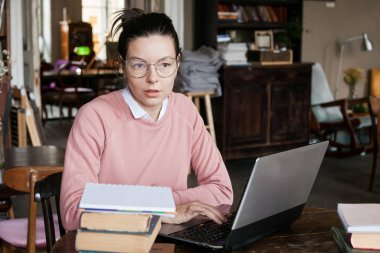 Sınava hazırlık. Gözlük ve pembe bir kazak çekici esmer öğrenci kız, hafif bir gülümseme ile bir dizüstü bilgisayar oturur.