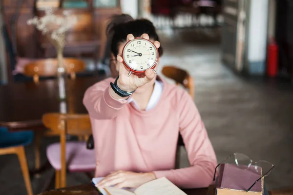 Prazo limite. Um despertador de close-up voltado para a moldura realizada por uma garota morena em um suéter rosa à mesa em uma biblioteca antiga . — Fotografia de Stock