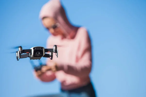 De pie con una rodilla doblada, la joven con capucha rosa y gafas oscuras controla el panel de control de vuelo del dron . —  Fotos de Stock