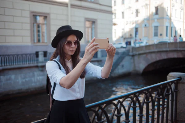 Turysta w mieście robi zdjęcie na smartfonie. Młoda kobieta w czarnym kapeluszu i biała koszula, — Zdjęcie stockowe