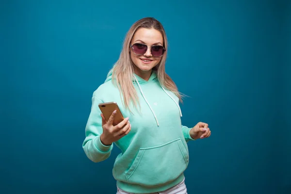 Stylový trendy hipster dívka v modré mikině s kapucí používá smartphone. — Stock fotografie