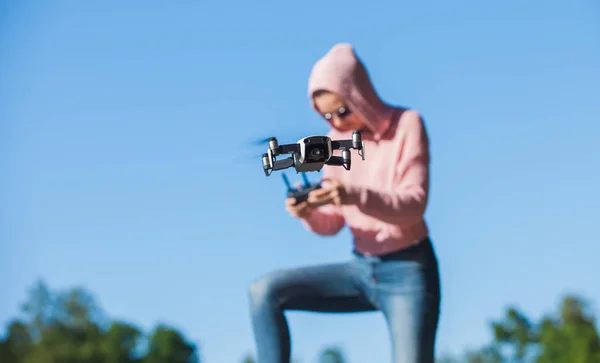 Debout avec un genou plié, une jeune femme avec un sweat à capuche rose et des lunettes sombres contrôle le panneau de commande de vol du drone . — Photo