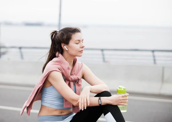 Athletic young woman in sports outfit after jogging, drinks water from green bottle, sitting on the parapet of bridge.