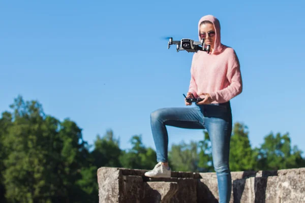 Une jeune femme avec un sweat à capuche rose et des lunettes de soleil contrôlées avec la télécommande drone vol dans la nature . — Photo