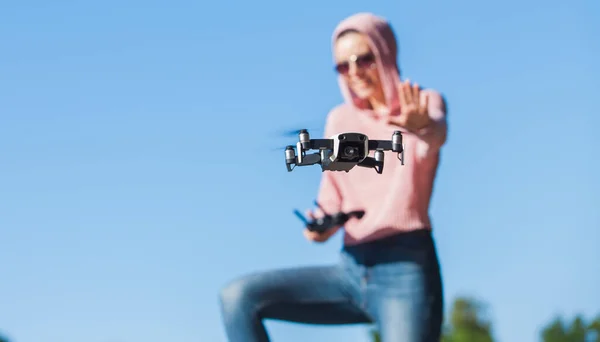 Standing with a bended knee, young woman in pink hoodie and dark glasses controls the drone\'s flight control panel.