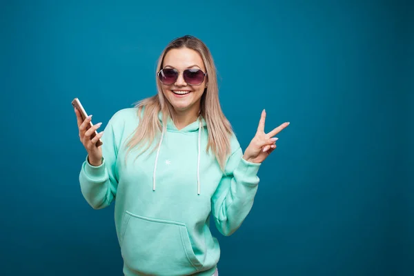 Stylový trendy hipster dívka v modré mikině s kapucí používá smartphone. — Stock fotografie