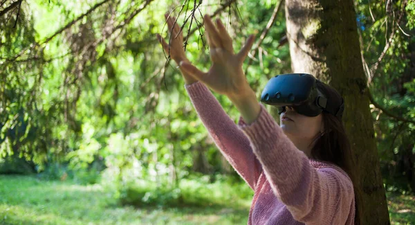 Virtual forest. A young woman among the trees, wearing virtual reality glasses, touches the boles and branches.