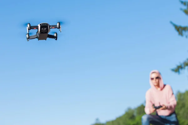 De pie con una rodilla doblada, la joven con capucha rosa y gafas oscuras controla el panel de control de vuelo del dron . —  Fotos de Stock