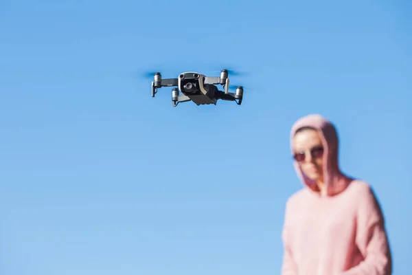 Standing with a bended knee, young woman in pink hoodie and dark glasses controls the drone's flight control panel.