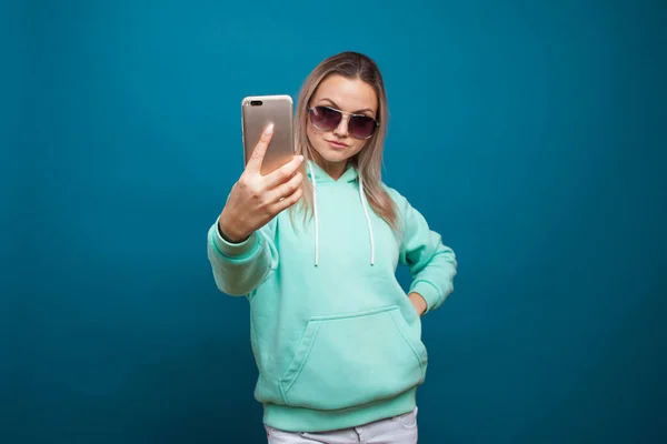 Elegante moderno hipster menina com capuz azul com o capuz usa um smartphone. — Fotografia de Stock